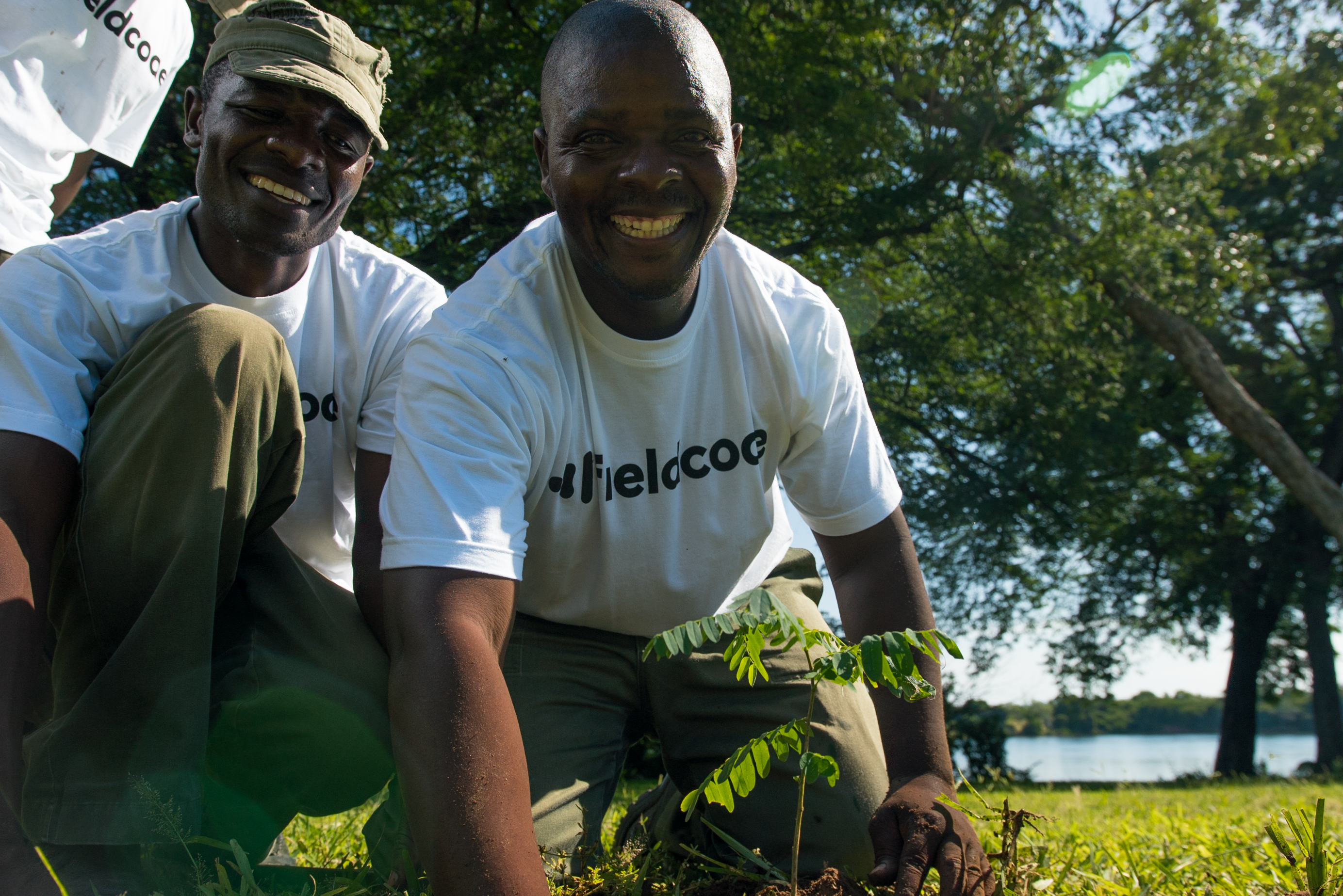 Two smiling men sit on the grass in front the river while planting a small tree in Lower Zambezi Valley in Zimbabwe Two smiling men sit on the grass in front the river while planting a small tree in Lower Zambezi Valley in Zimbabwe